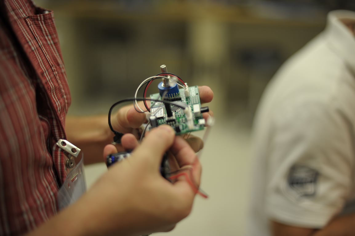 Hands examine a green Humbo circuit board with a blue rotary control and connected wires in the workshop.