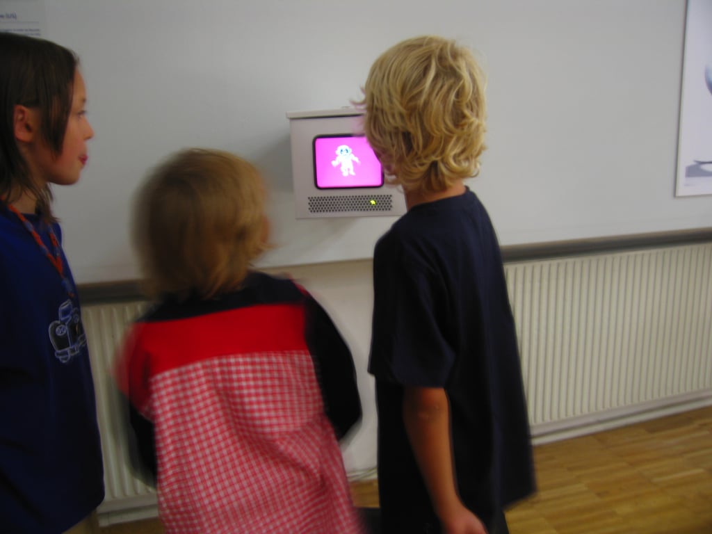 Three children are watching a small screen displaying a cartoon character on a pink background.