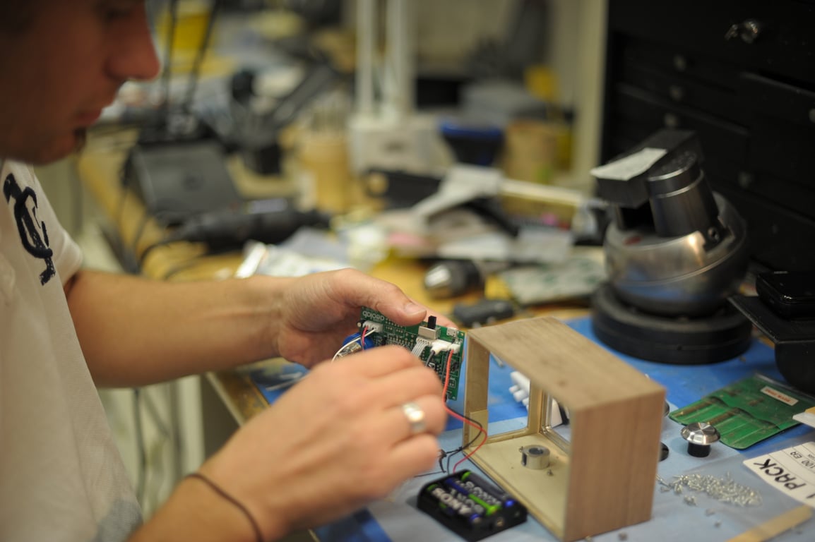 A person handles a wired circuit board beside an open wooden Humbo enclosure and battery holder on a workbench.