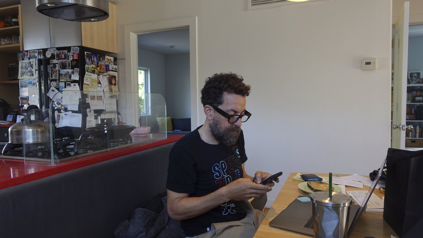 Julian at the kitchen table with a phone, laptop, papers and breakfast.