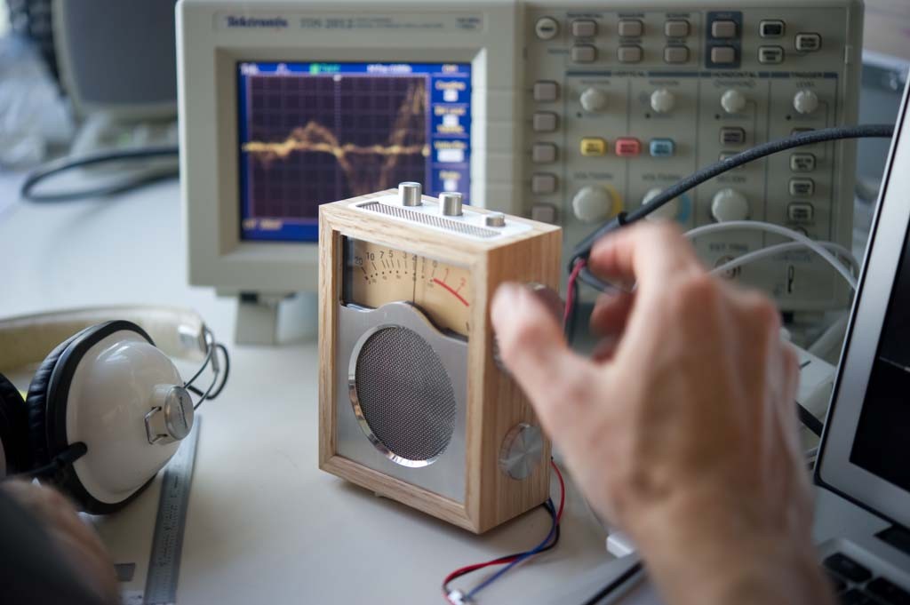 A hand adjusts the side dial of the assembled wooden Humbo radio while an oscilloscope displays a waveform behind it and headphones rest on the bench.