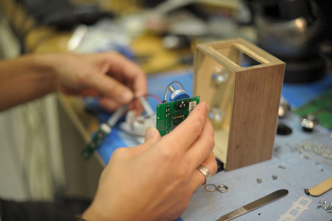 Hands hold the underside of a green Humbo circuit board beside an upright wooden enclosure and loose metal hardware.