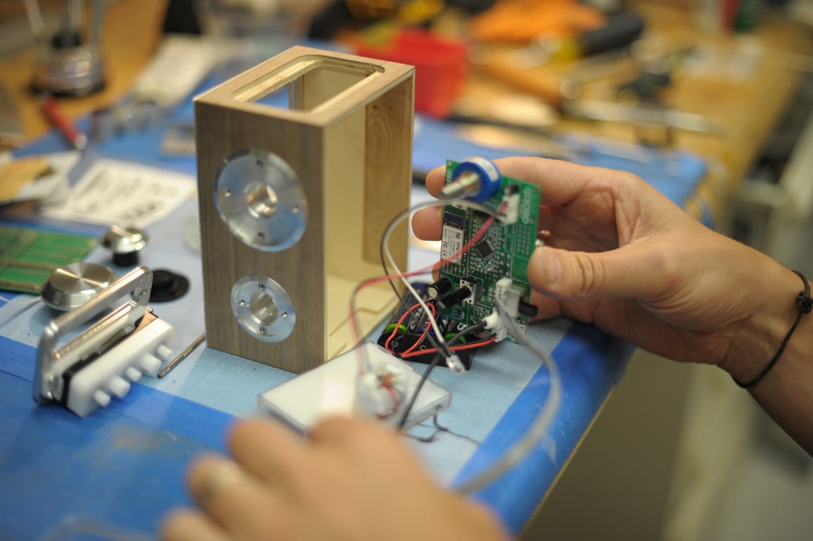 Hands hold the Humbo circuit board beside an upright wooden enclosure with two metal side fittings and a loose wired panel.