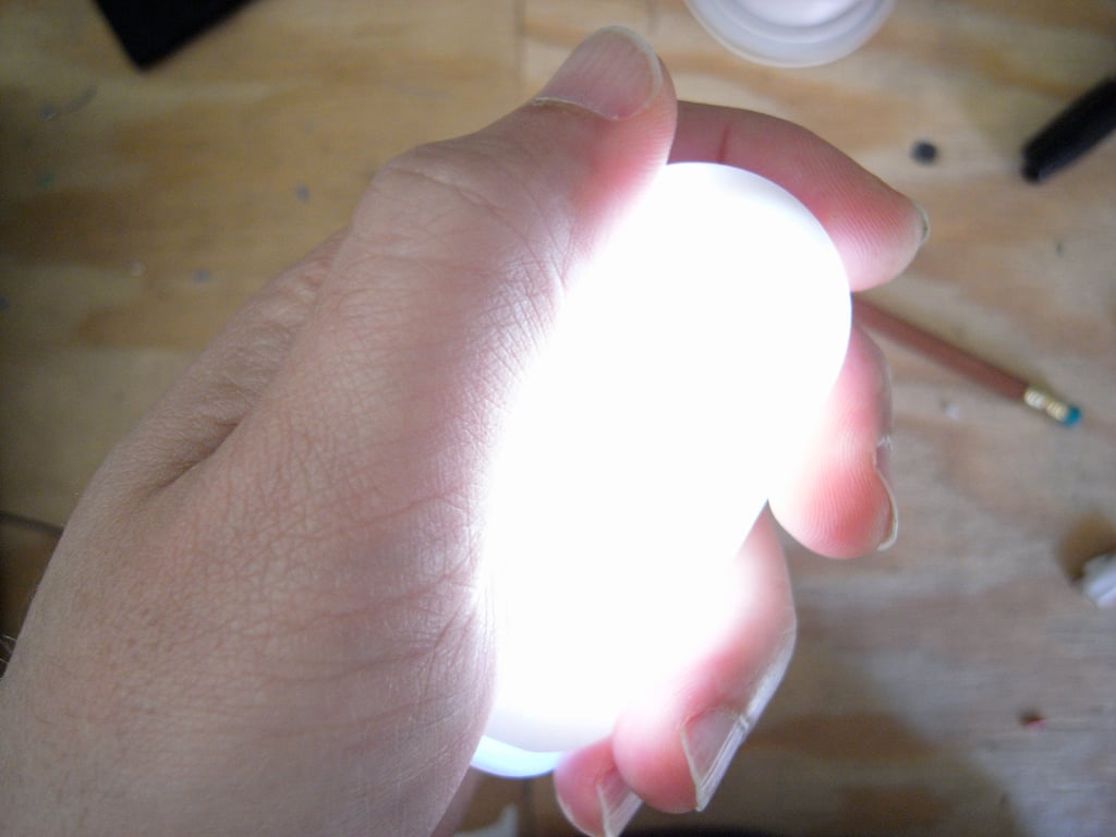 A hand wrapped around the smaller Kombolói enclosure glowing bright white above a wooden workbench.