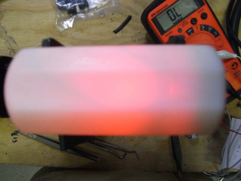 White Kombolói enclosure glowing red on a wooden workbench beside an orange multimeter.