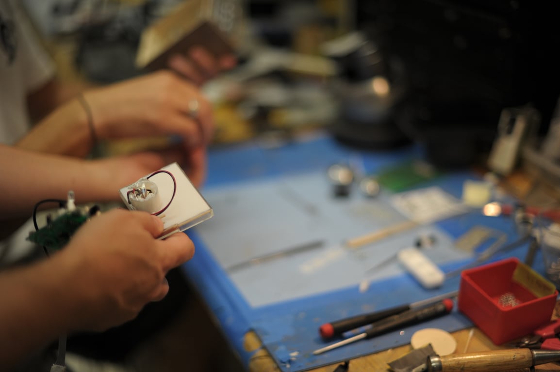 Hands hold a small wired panel in the foreground while another person works at the same tool-covered bench.