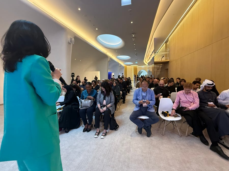 A woman in a turquoise suit speaks to an audience seated in a modern conference room.