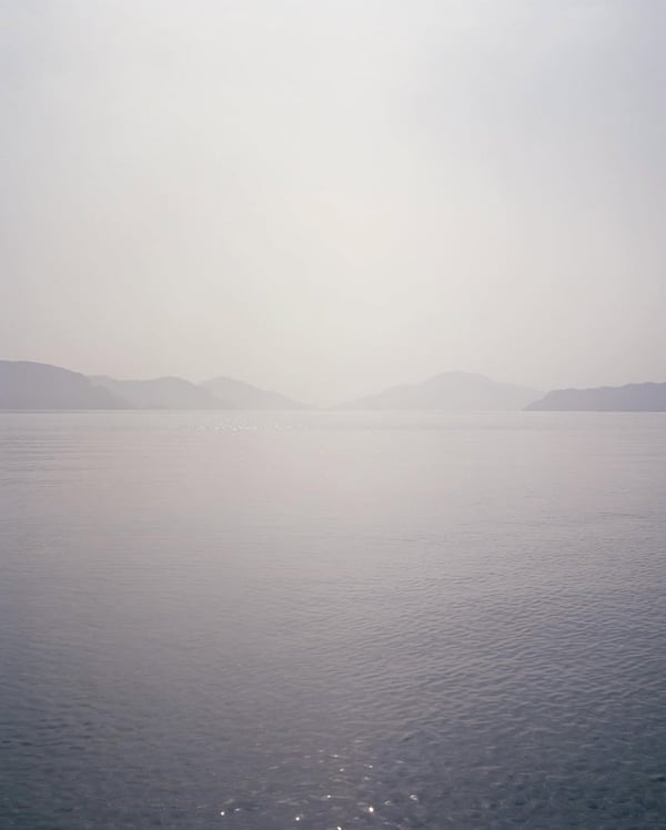 Calm sea with distant hazy mountains under a bright, overcast sky.
