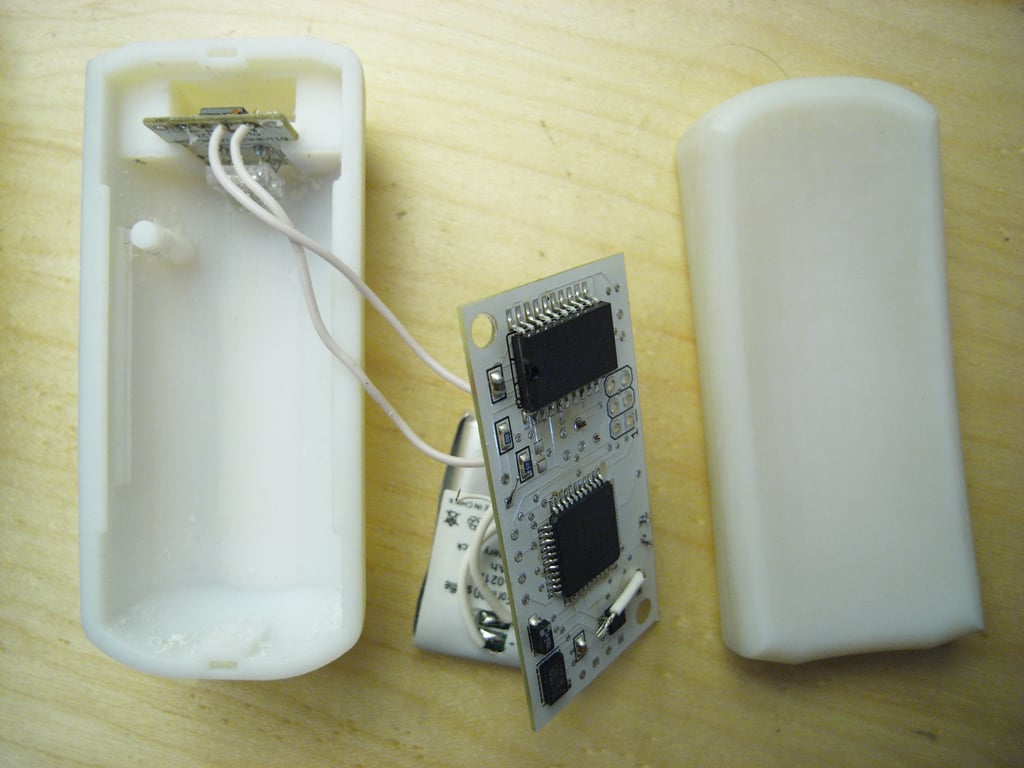 Opened white Kombolói enclosure with its exposed printed circuit board, white wiring, and battery components on a wooden table.