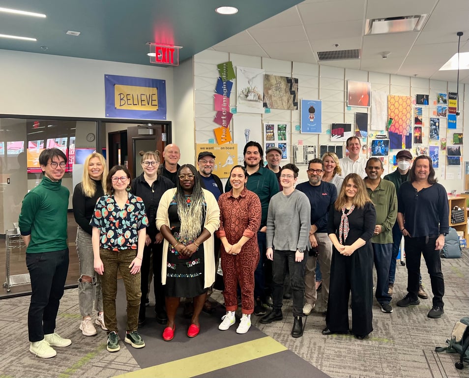 Group of people collaborating in a bright room with tables, whiteboards, and colorful wall art.