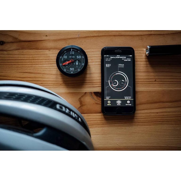 An OMATA One instrument and an iPhone displaying the ride dashboard arranged with a bicycle helmet and handlebar component on a wooden work surface.