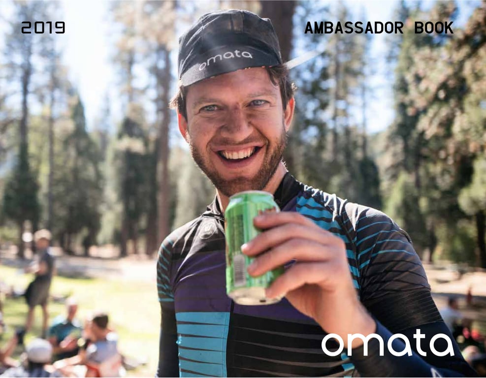 April 2019 OMATA Ambassador Book cover showing a smiling cyclist holding a green drink at an outdoor gathering.