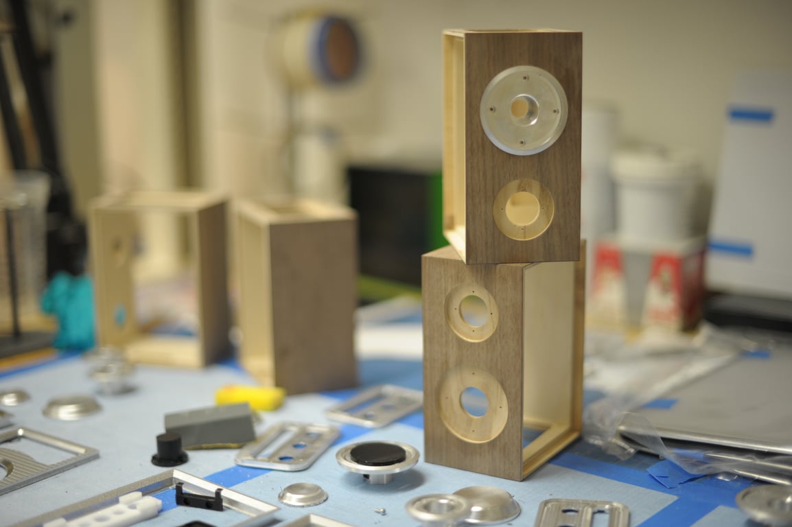 Two wooden Humbo enclosures stacked on a workbench, showing circular side recesses, a fitted metal disc, and loose metal parts nearby.