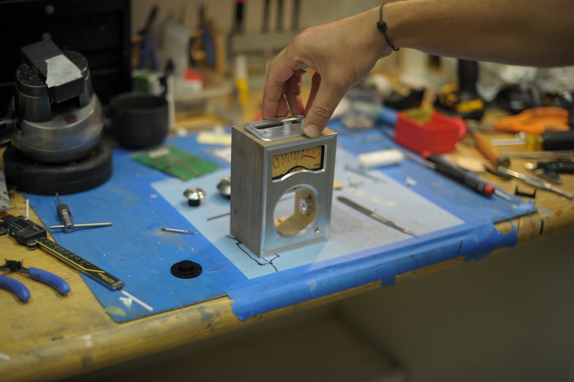 A hand fits a metal top panel onto the wooden Humbo enclosure, with the front meter installed and the speaker opening empty.