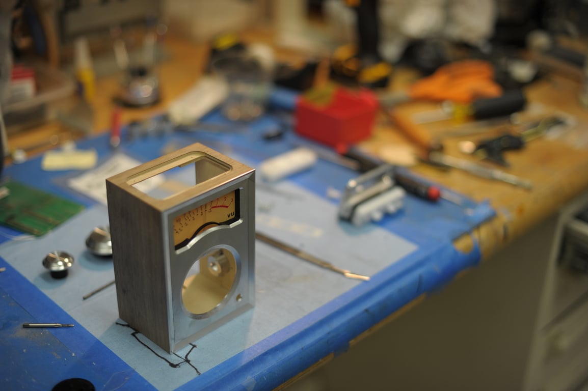 Wooden Humbo enclosure standing on a workbench with its metal front and analog meter fitted, leaving the top and speaker opening empty.