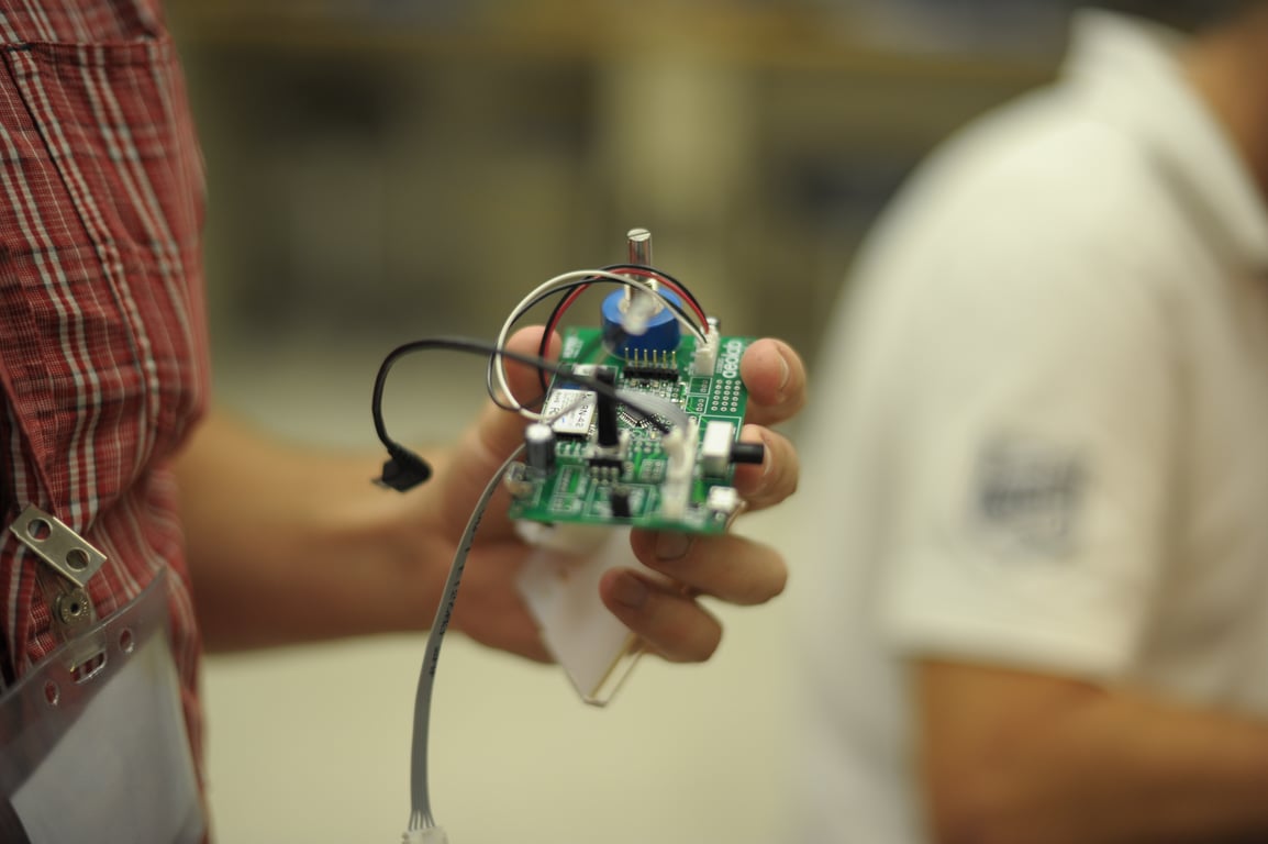 A hand holds the green Humbo circuit board with its blue rotary control, connectors, and attached wires.