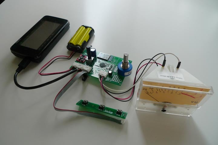 Humbo Personal Radio Station working prototype on a workbench, with an N900, yellow battery pack, custom circuit board, blue tuning dial, switch assembly, and analog VU meter.