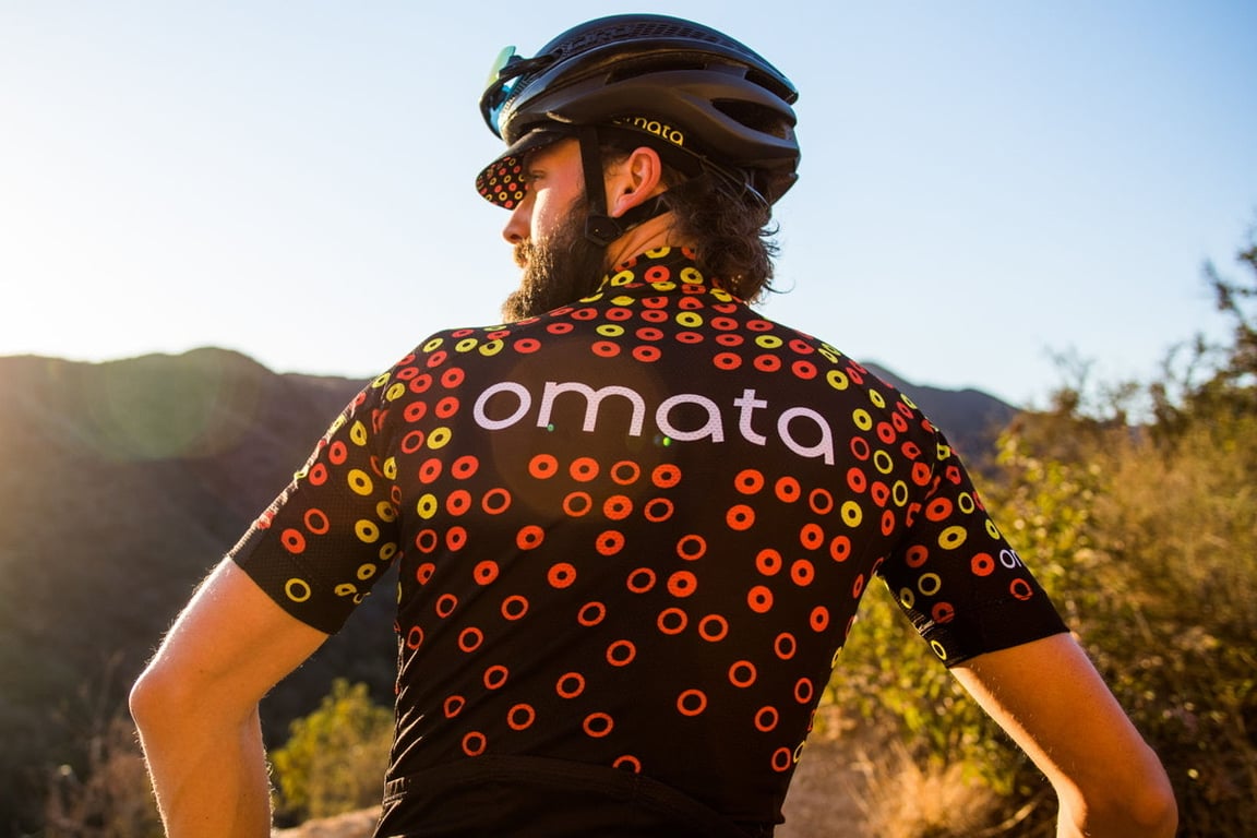 Cyclist seen from behind wearing the black OMATA cycling jersey with red, orange, and yellow ring patterns.