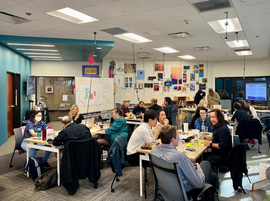 Group of people collaborating in a bright room with tables, whiteboards, and colorful wall art.