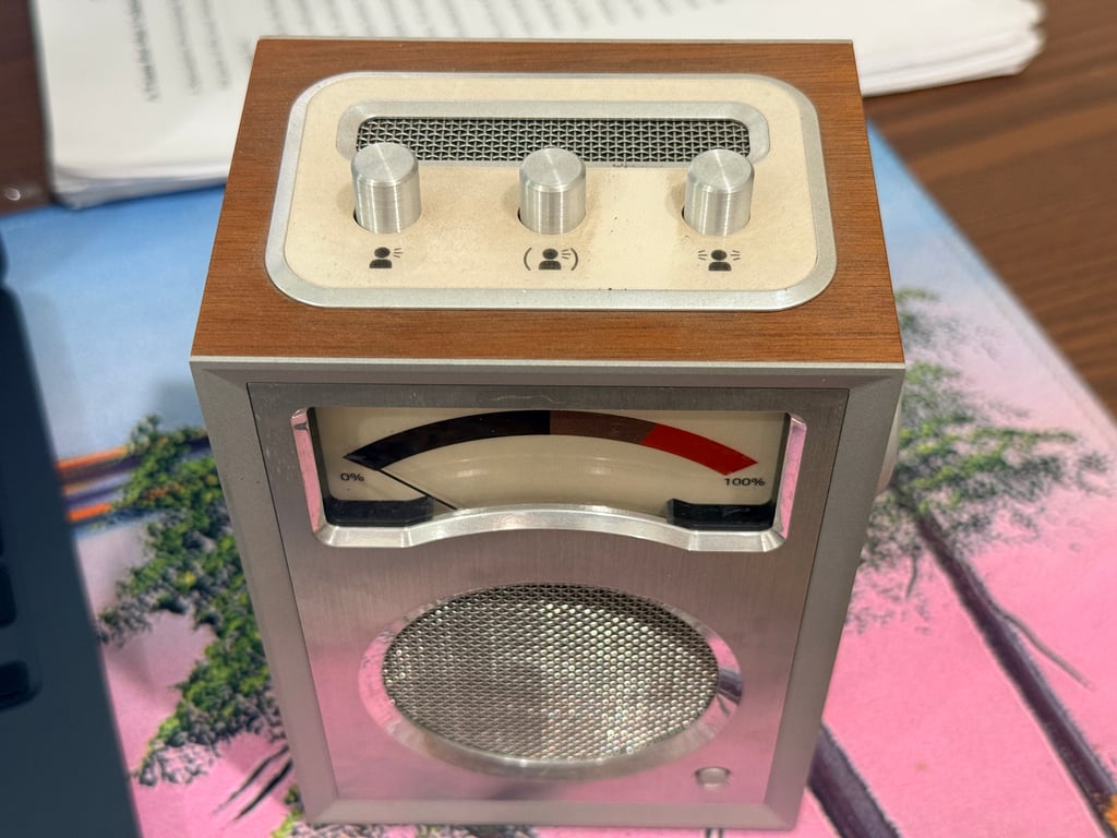 Humbo viewed from above and in front, showing three small metal top controls, a mesh opening, a shaped analog meter window, and a circular speaker grille.
