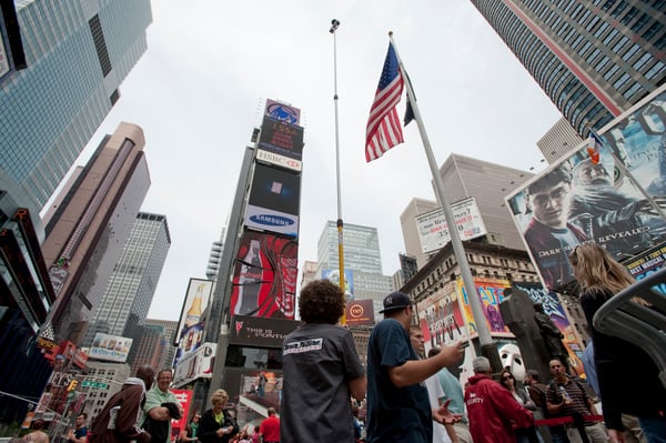 Crowd in Times Square with tall buildings, billboards, and an American flag on a cloudy day.