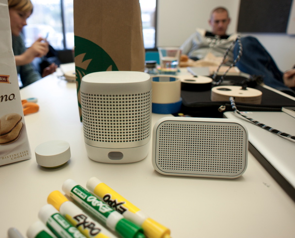 White cylindrical and rectangular speaker prototypes on a studio table with markers and design materials.