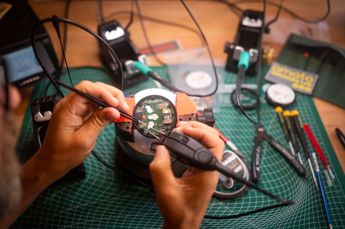 Hands working on a small OMATA instrument at a bench with soldering tools and components.