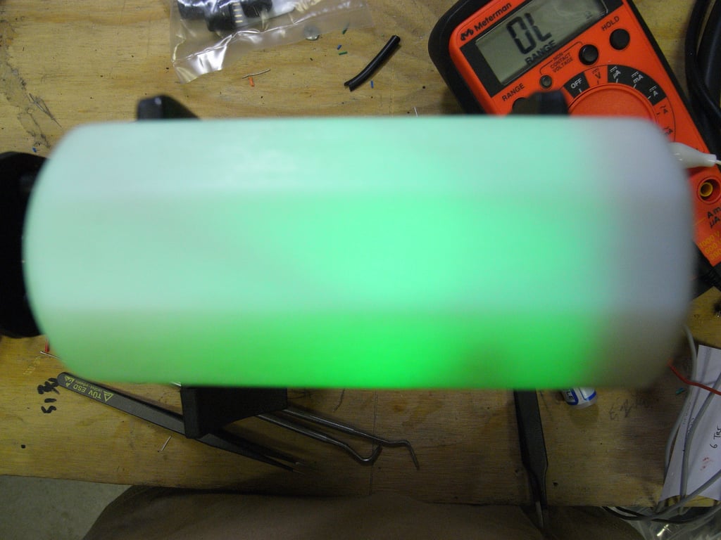 White Kombolói enclosure glowing green on a wooden workbench beside an orange multimeter.