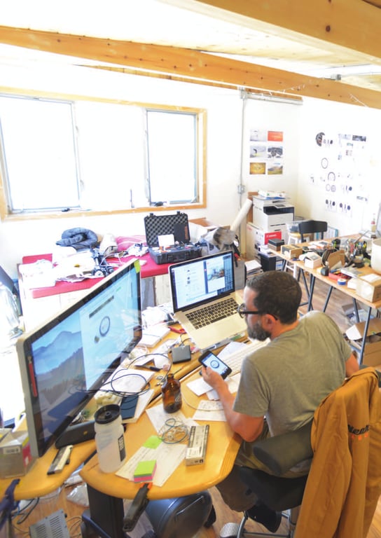 Overhead view of Julian Bleecker working at a desk with a laptop, monitor, and tablet in his converted garage-studio.