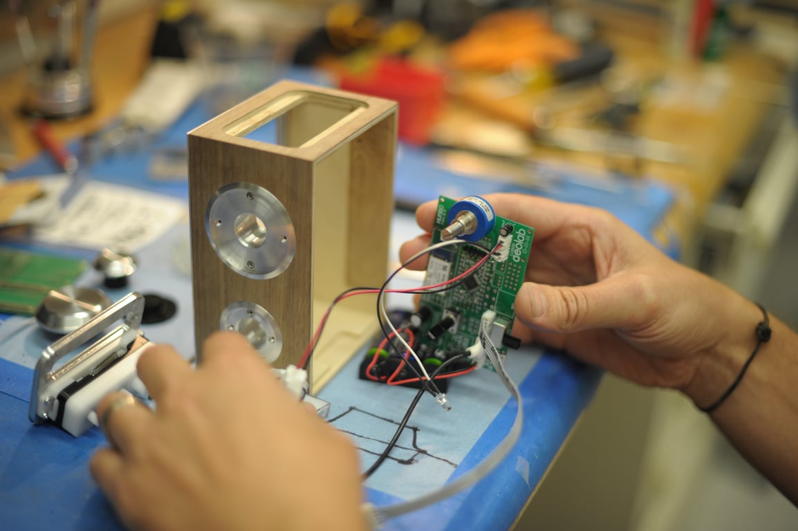 A hand holds the Humbo circuit board and blue rotary control next to a wooden enclosure with circular metal side fittings.