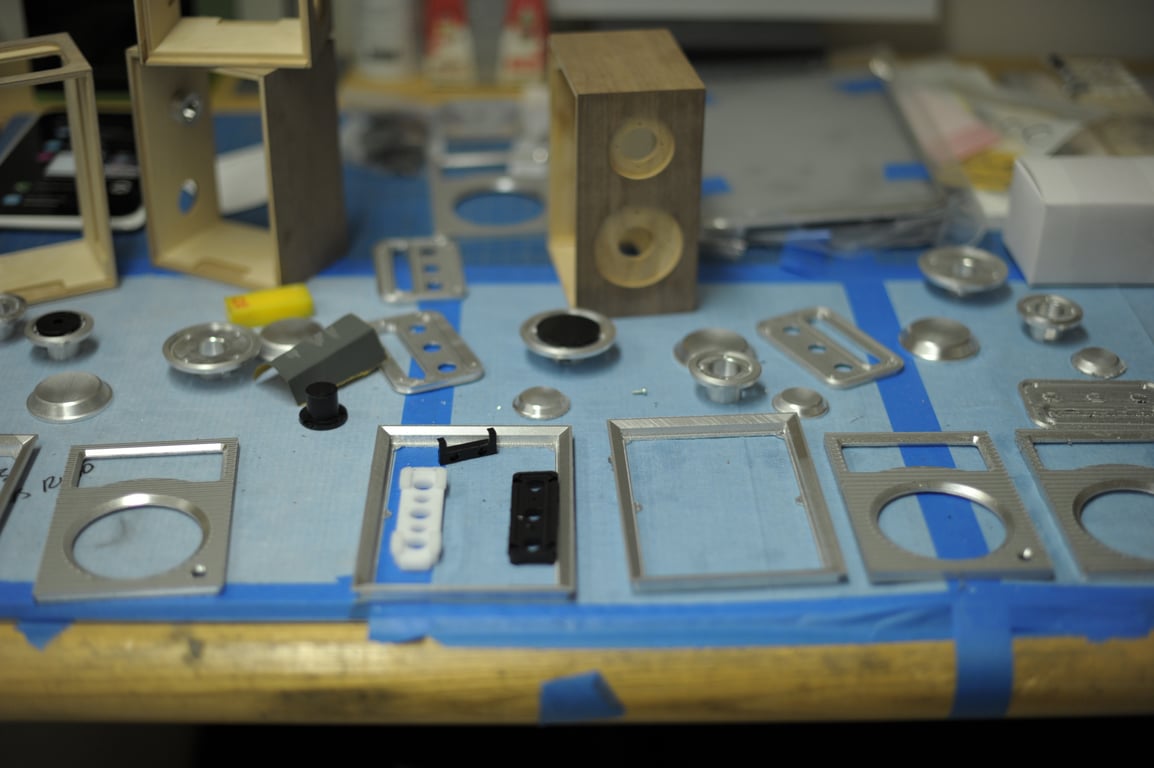 Wooden Humbo enclosures, machined metal faceplates, knobs, and switch panels arranged across a blue-covered workbench.