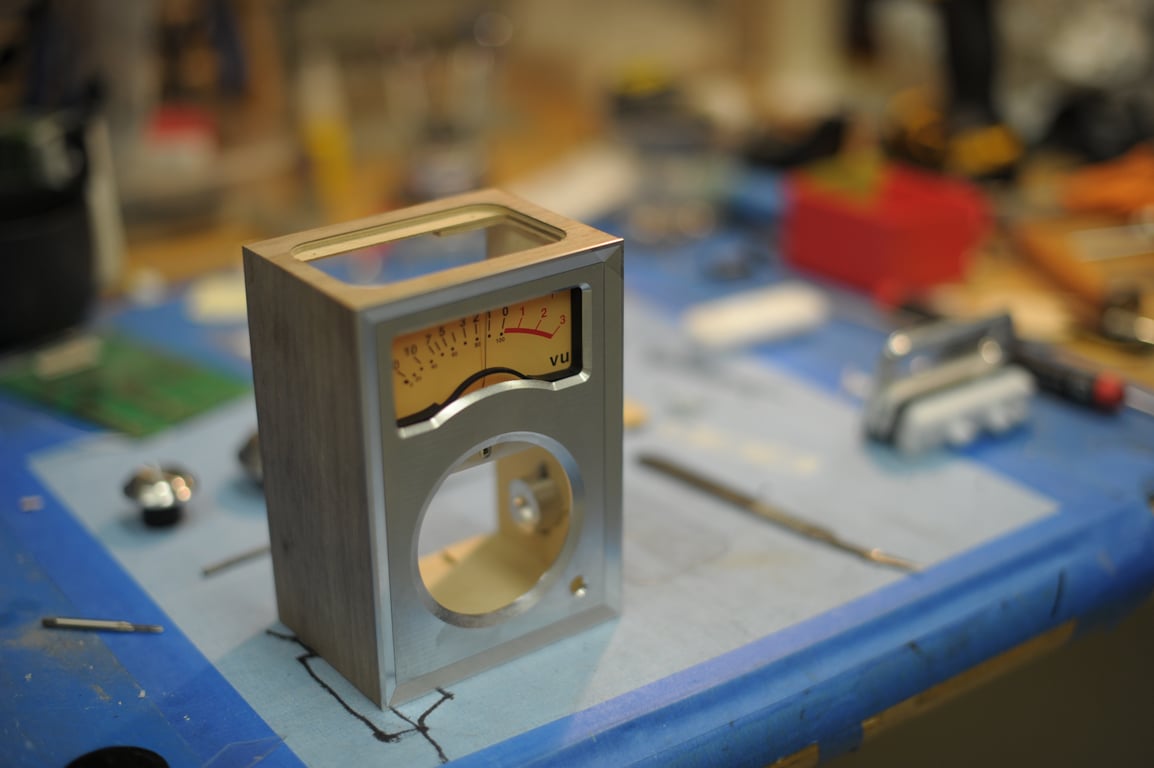 Three-quarter view of a wooden Humbo enclosure with a fitted metal front, analog VU meter, and open circular speaker aperture.
