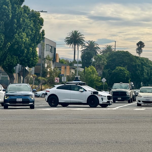 A white autonomous vehicle with a sleek, modern design and black wheels is positioned blocking an intersection in an urban setting.