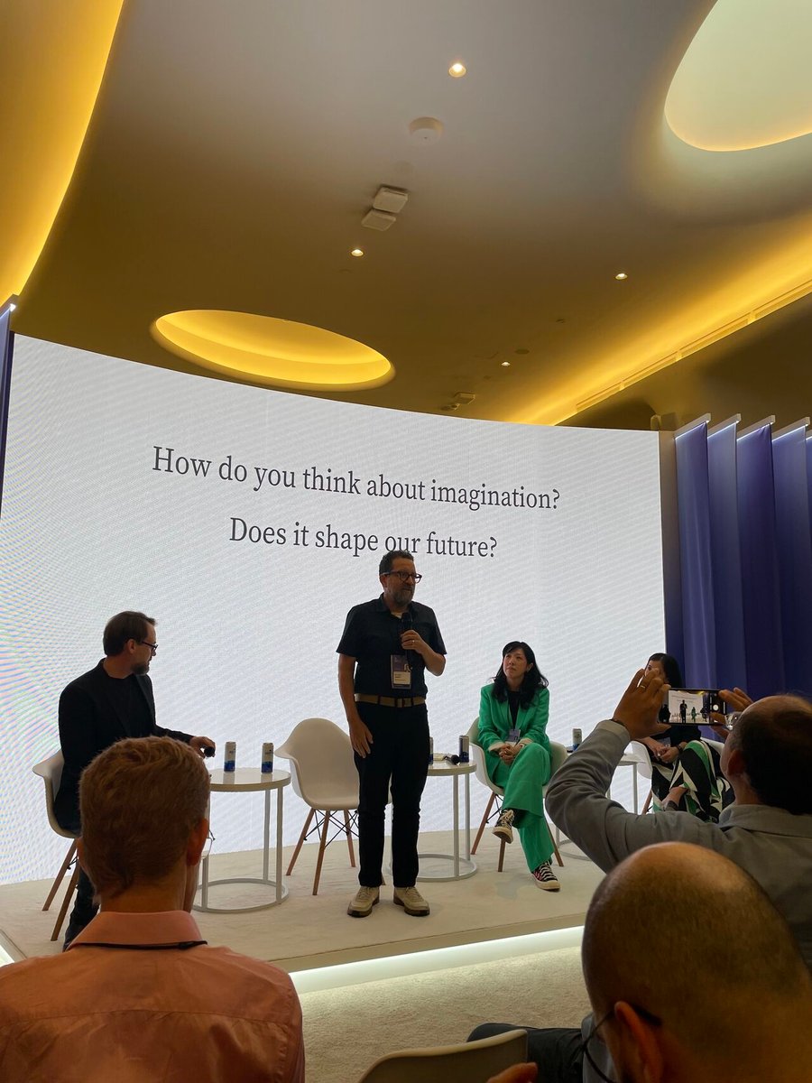 Panel discussion with three people on stage, audience in foreground, text on screen about imagination and future.