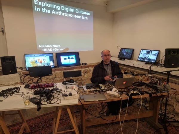 Nicolas Nova sits at a table with multiple screens and audio equipment, presenting on digital cultures.
