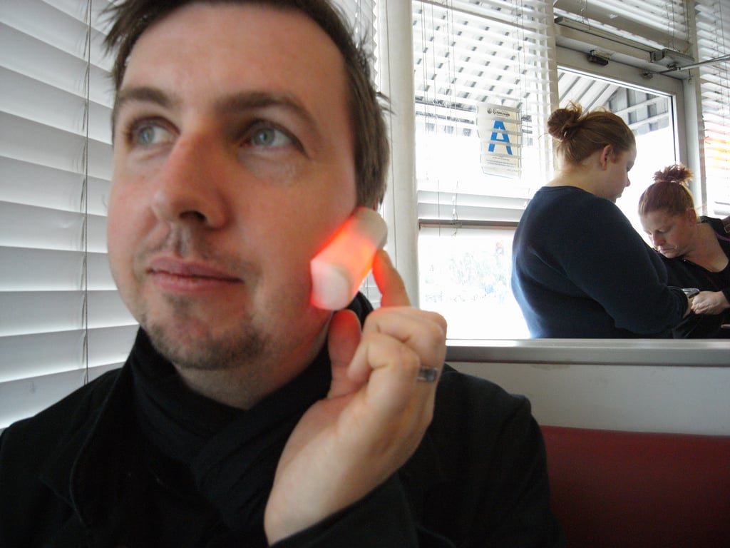A man holding the red-glowing Kombolói against his cheek while seated in a diner.