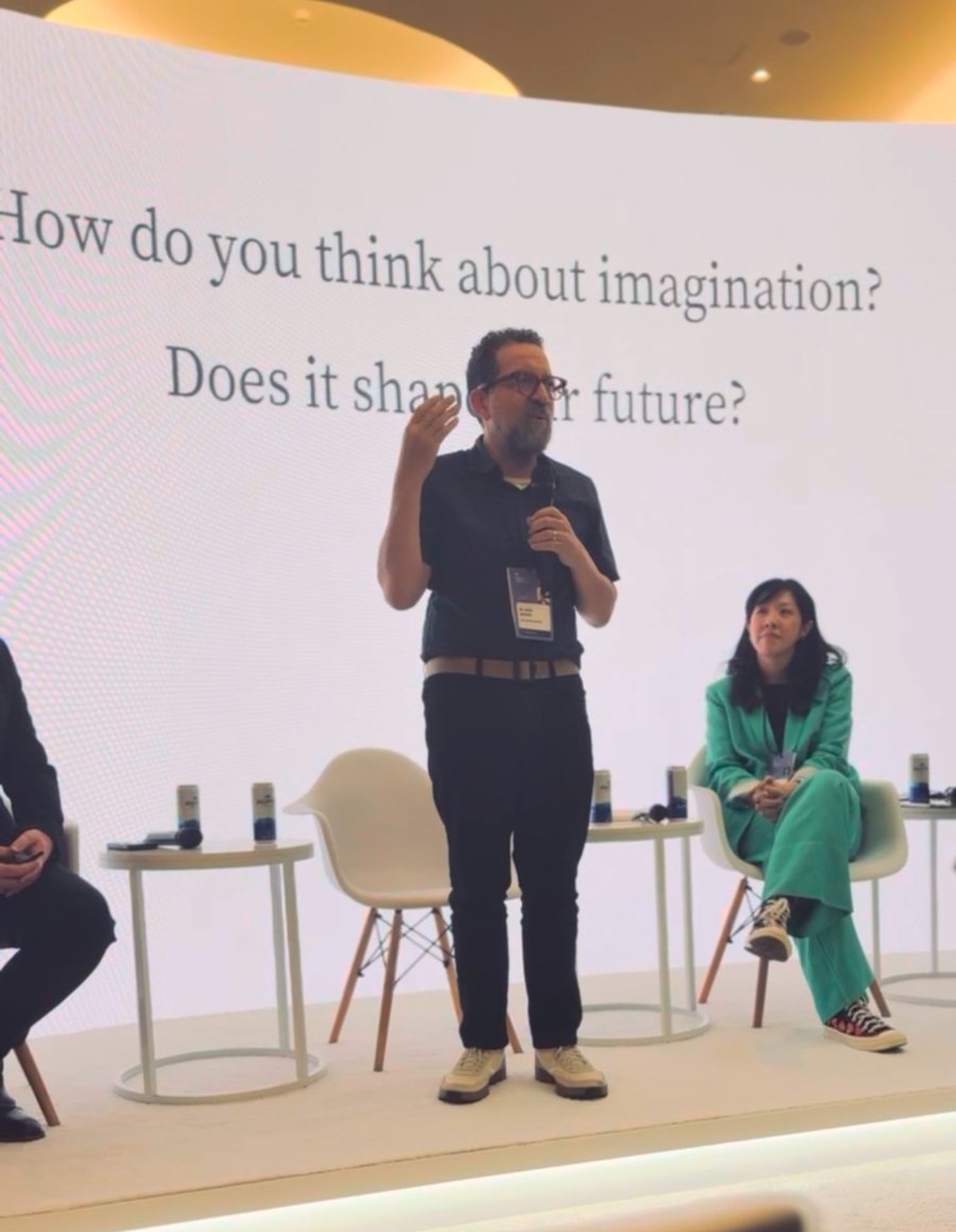 Man speaking on stage with a microphone, text behind him reads, "How do you think about imagination? Does it shape our future?"