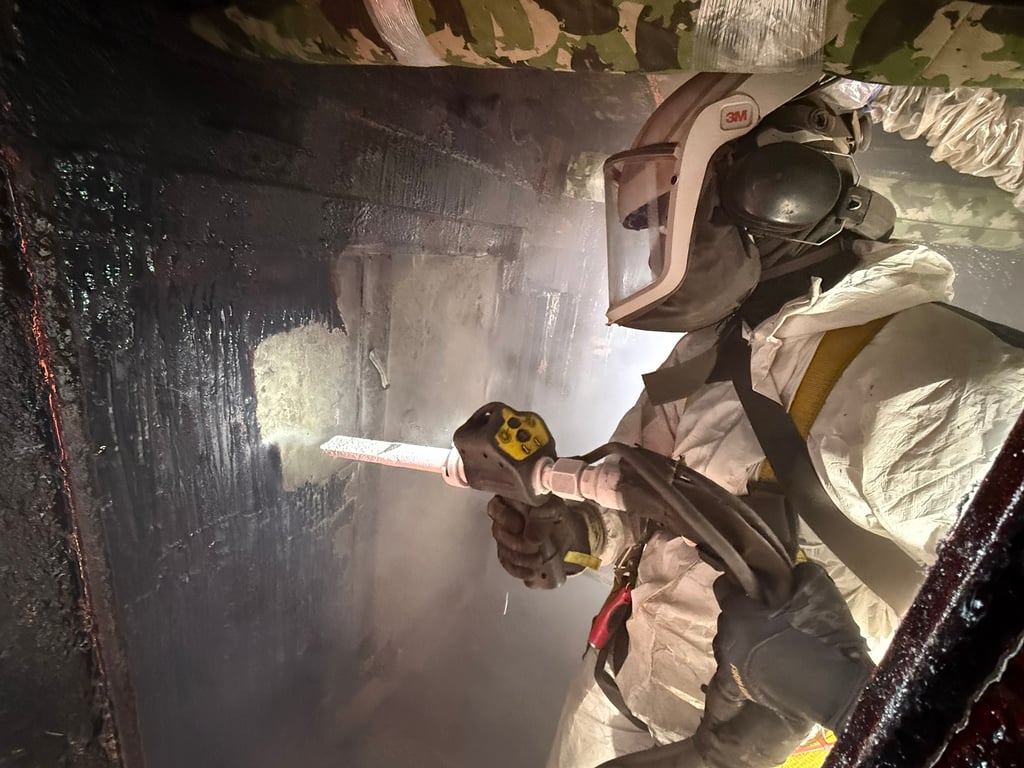 Technician in full OSHA-compliant PPE and 3M respirator blasting soot from a fire-damaged surface