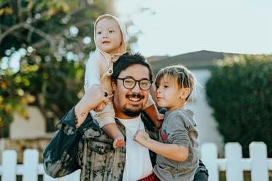 A man holding two young children, one on each arm. The man is smiling and wearing glasses and a denim jacket. The children are dressed in casual clothing, and they all appear happy. They are standing outside with a white fence in the background.