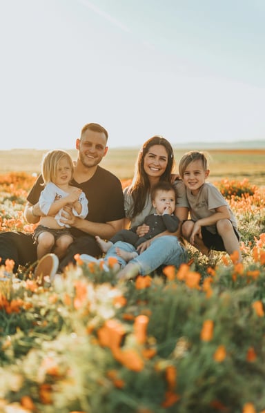 A family sitting in a field of orange flowers. The father is holding a child on his lap, the mother is holding a baby, and another child is sitting close to them. They are all smiling and enjoying the sunny day.