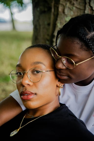 Two women wearing glasses, with one woman resting her head on the other's shoulder. They are outside near a tree, looking content and connected. The woman in front is wearing a black top and gold jewelry, while the woman behind her is wearing a white shirt.