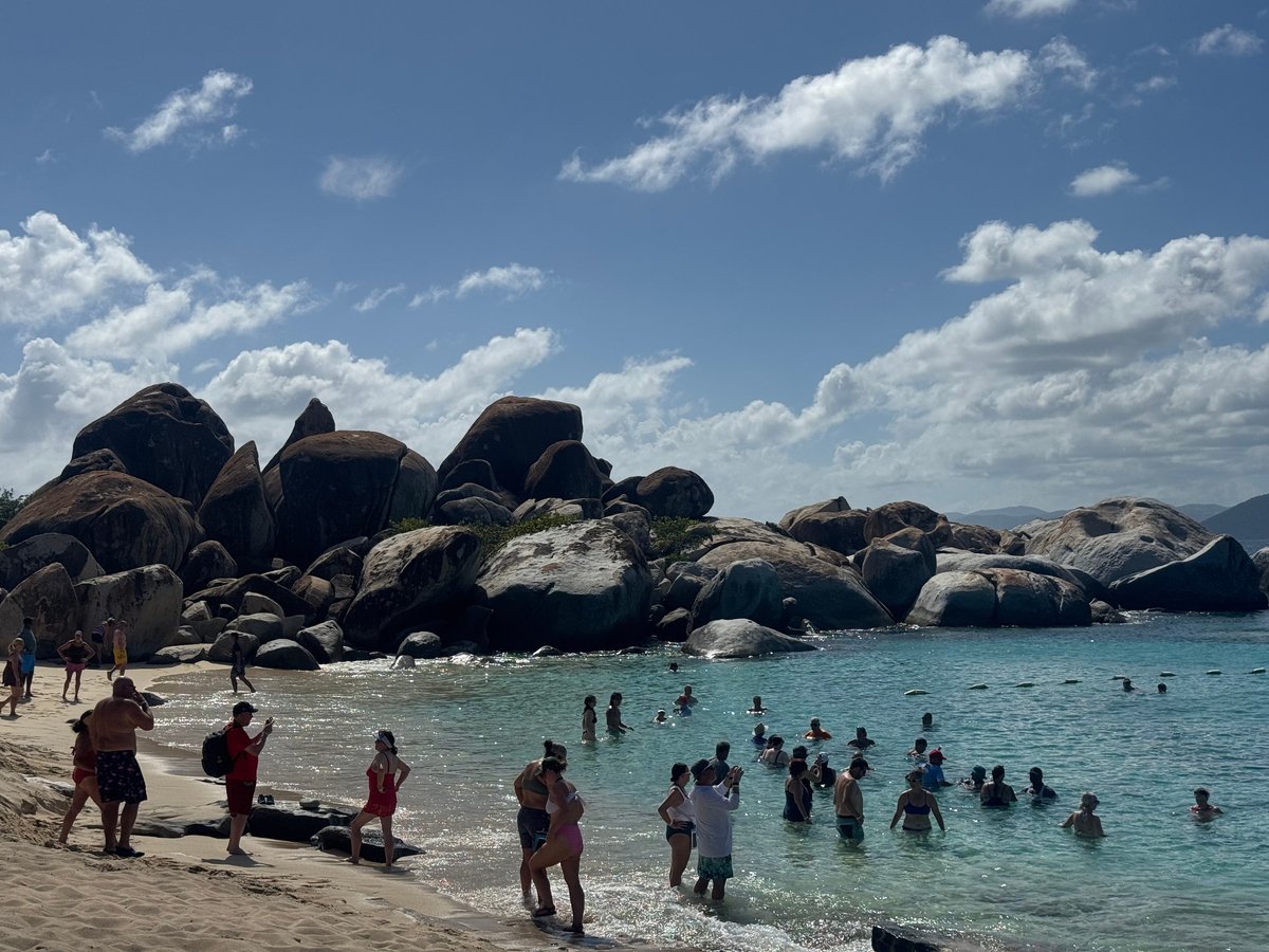 Families and tourists swimming and wading in turquoise water near a sandy beach with massive rounded granite boulders under a blue sky with white clouds.