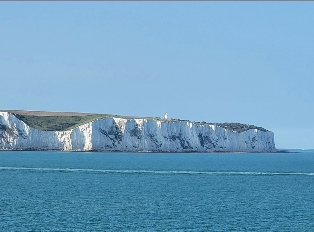 Dramatic white chalk cliffs rise from turquoise waters under clear blue sky, with green coastal grassland atop.