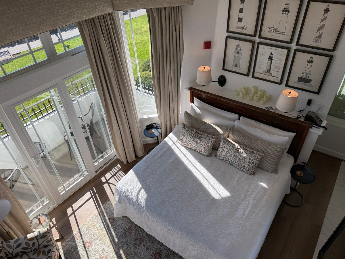 Bright, modern bedroom with a queen bed, green coastal views through floor-to-ceiling glass doors, lighthouse artwork, and candles on the nightstand.