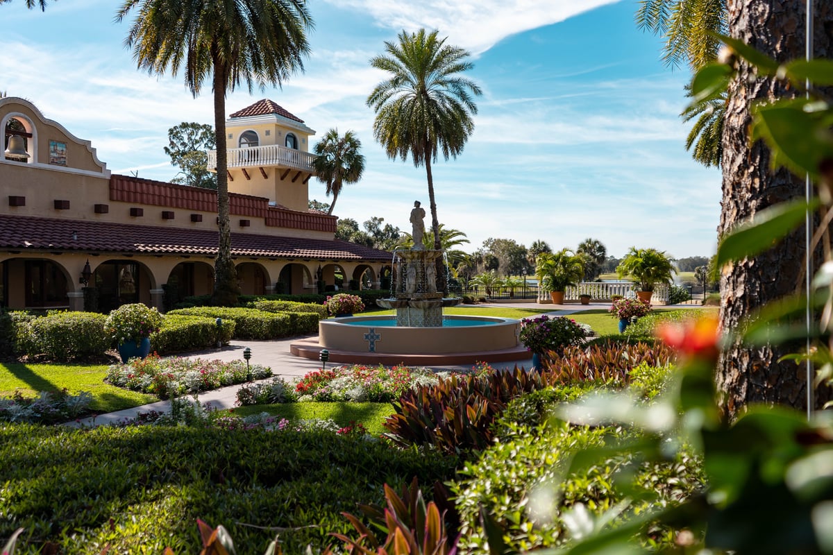 A serene courtyard with manicured gardens, a blue fountain, colonial architecture with arched portico, and palm trees under clear blue sky.