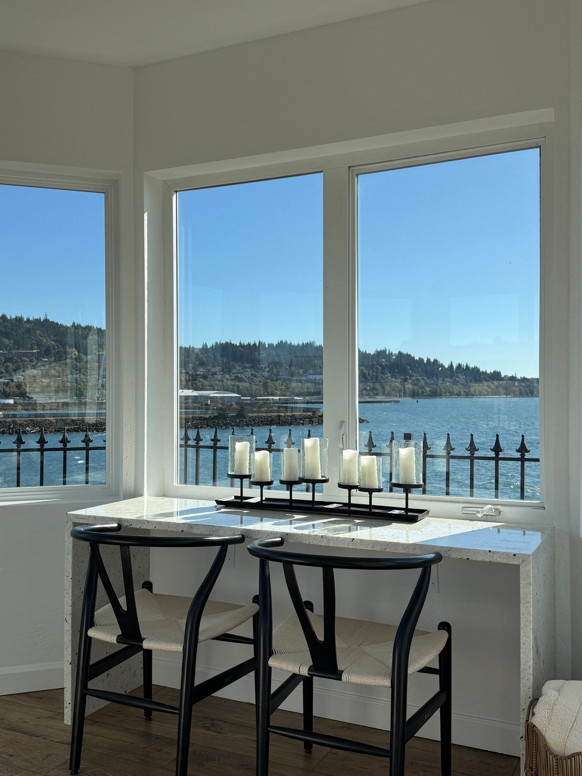 Two black chairs at a white desk facing floor-to-ceiling windows overlooking a waterfront with forested shores and a decorative candelabra.