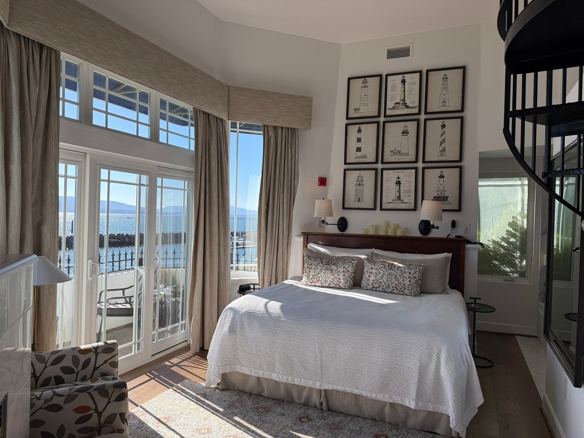 A bright, modern coastal bedroom with ocean views through glass doors, a queen bed with neutral bedding, lighthouse artwork, and a spiral staircase visible on the right.