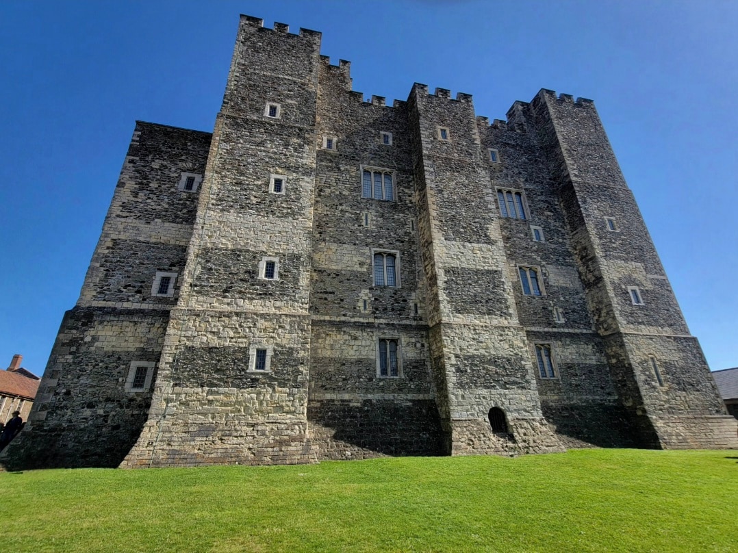 A grand medieval stone castle with multiple crenellated towers stands against a clear blue sky, viewed from a manicured green lawn.