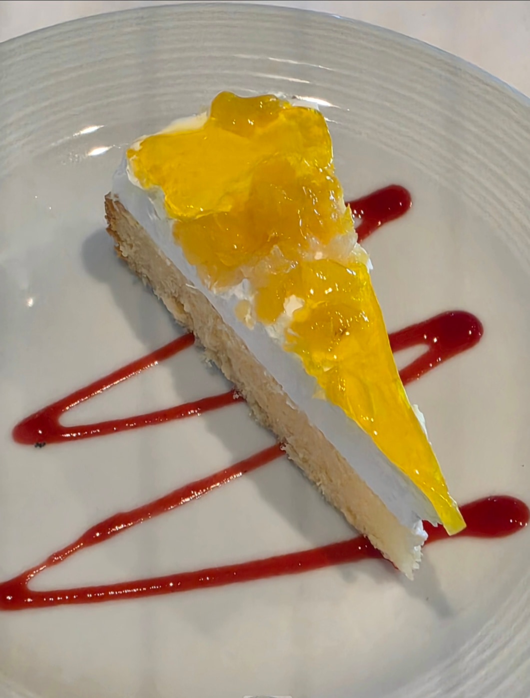 A plated dessert featuring a layered cake with white cream and yellow fruit topping, garnished with red sauce drizzle on a white dish.