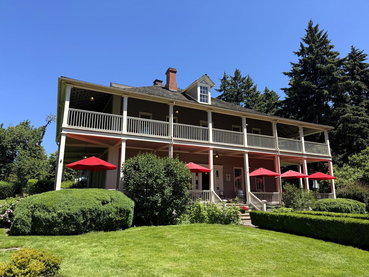 A stately colonial-style inn with white railings and red umbrellas sits amid manicured green lawns and evergreen trees under clear blue sky.